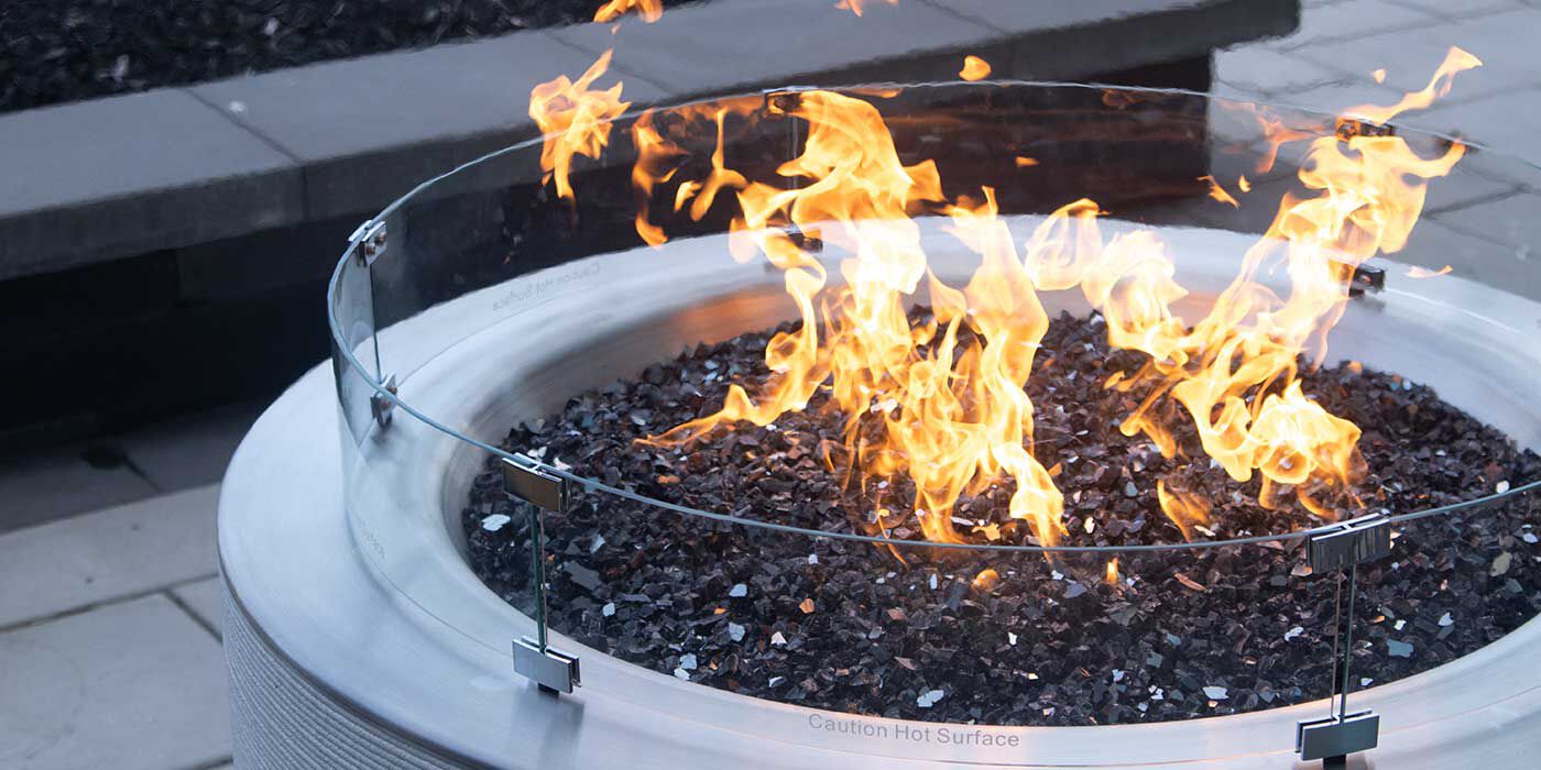 The ReForm Stack Gas Fire Pit burning outside with a glass fire pit wind guard attached around the opening.