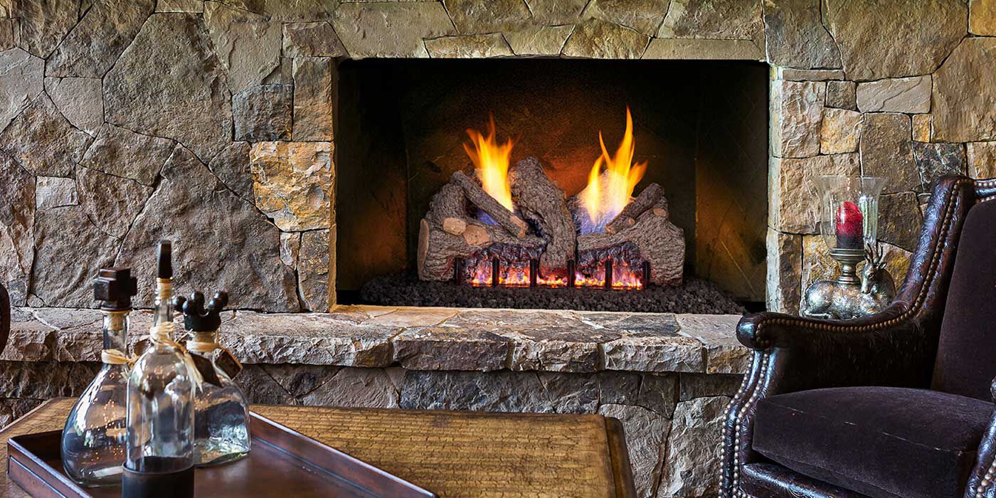 A rustic, traditional living room with a velvet decorative chair and wooden accents, along with a large, stone fireplace, a wooden mantel, and a Ventless gas log set.