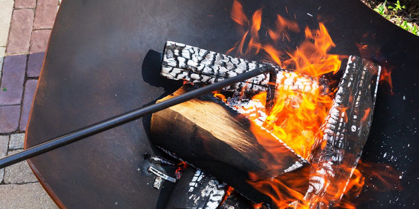 A close-up of a wood fire pit burner and someone moving the logs around with a poker