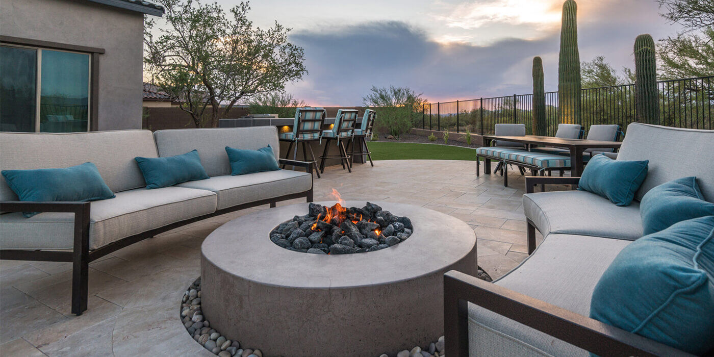 An outdoor patio space with a large, round concrete fire pit and two sectional outdoor couches