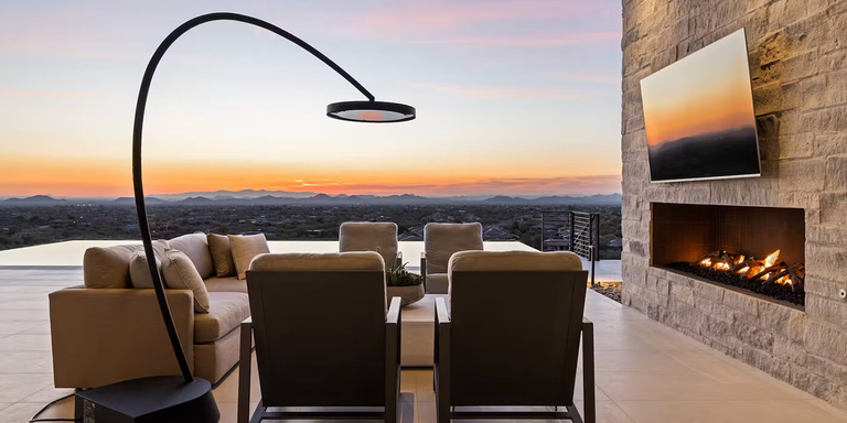 A photograph from a patio looking out at an infinity pool and a sprawling valley landscape during a colorful sunset. The outdoor living space has a sectional sofa and lounge chairs. To the left is a very large, black C-shaped arc floor lamp, and to the right, a television is mounted on a textured stone wall above an outdoor fireplace.