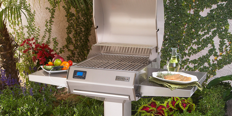 A Fire Magic electric grill on a patio featuring a digital display, flanked by side shelves holding fresh vegetables, seasoned fish, and olive oil.