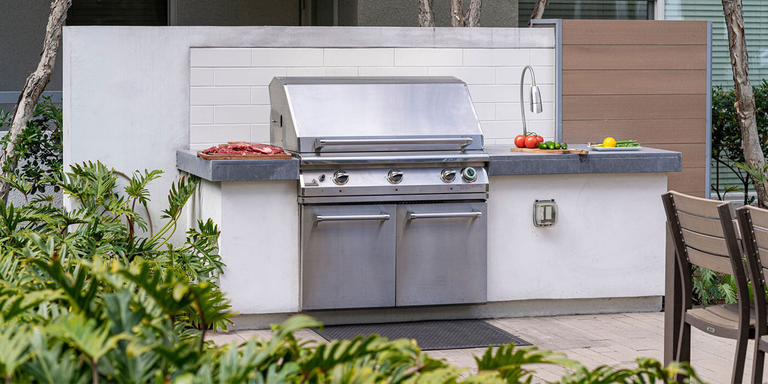 A contemporary outdoor kitchen in a patio setting with a stainless steel built-in grill and concrete countertops. A variety of grilling and dining ingredients, like steak, produce, and tools, are visible.