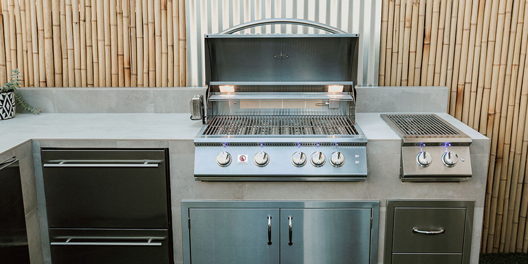 A built-in stainless steel grill with an open hood and internal lighting, integrated into a grey stone countertop. The setup includes a matching double-burner side station to the right and stainless steel storage drawers below. The background features a bamboo privacy fence and a corrugated metal accent panel.