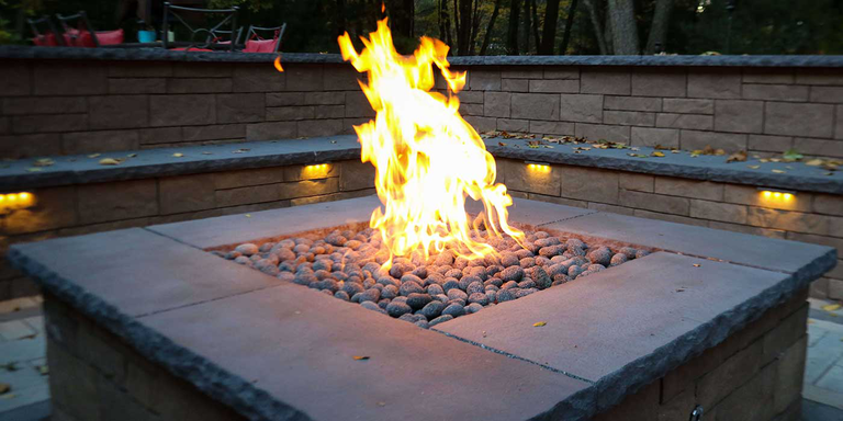A large, square, custom-built gas fire pit finished with natural stone