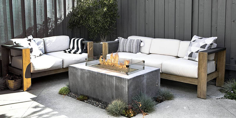 An outdoor living space with two couches and a concrete, rectangular gas fire pit with a glass wind guard 