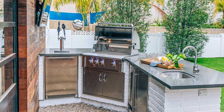 A detailed shot of a modern L-shaped outdoor kitchen with stainless steel appliances, a grey countertop, and white brick bases in a backyard setting.